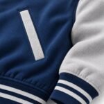 Close-up of varsity jacket shoulder seam with visible topstitching, silver-tone snap and navy-and-white striped rib collar
