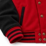 Close-up of topstitched black welt pocket, brass-tone snaps on the waistband tab and red and black striped rib cuff on a varsity jacket